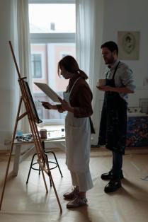 Two contemporary figurative artists in a sunlit studio—one painting at an easel, the other observing with a cup in hand.