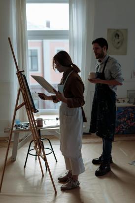 Two contemporary figurative artists in a sunlit studio—one painting at an easel, the other observing with a cup in hand.