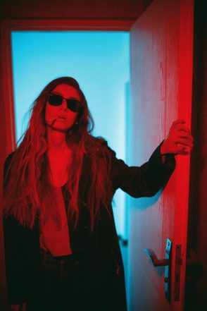 Woman with long blonde hair and sunglasses standing in a doorway with red and blue lighting