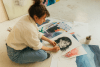 A female artist sits on the ground looking through a collection of drawings