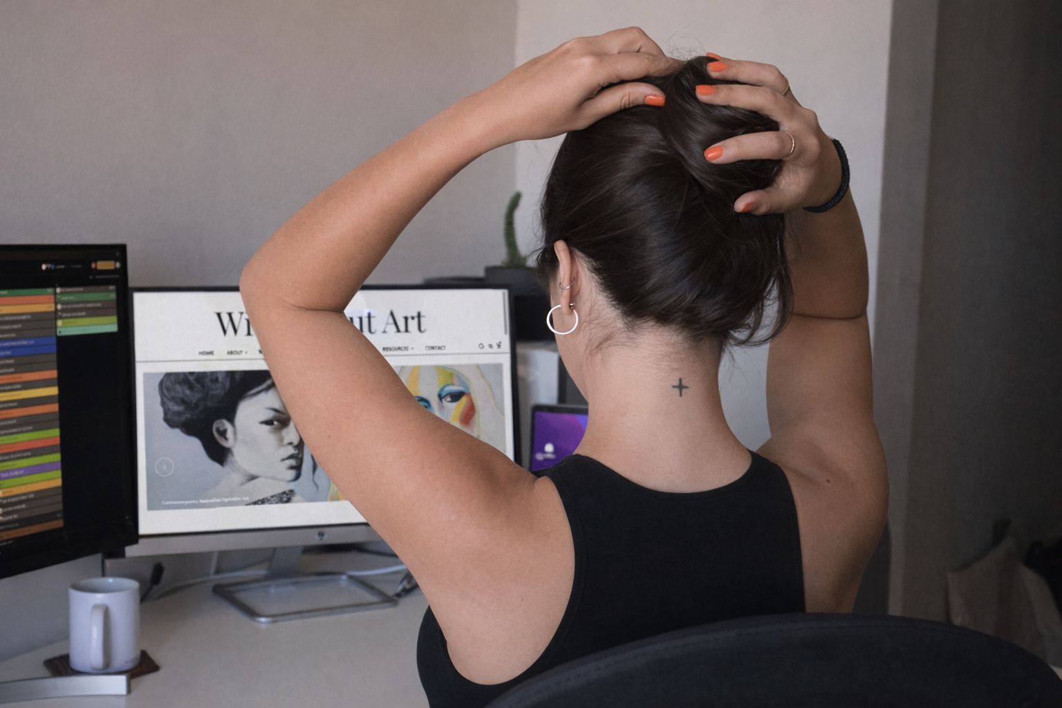 A woman with dark hair tied up sits at her desk, gathering her hair into a bun while looking at her computer screen. The monitor displays the *Wilde About Art* website, showing two portrait paintings — one monochrome and one brightly coloured — under the heading “Contemporary Australian Figurative Art.” She wears a black sleeveless top, has orange nail polish, and a small cross tattoo on the back of her neck. The workspace includes a second monitor, a white mug, and a small plant, creating a modern, creative studio atmosphere.