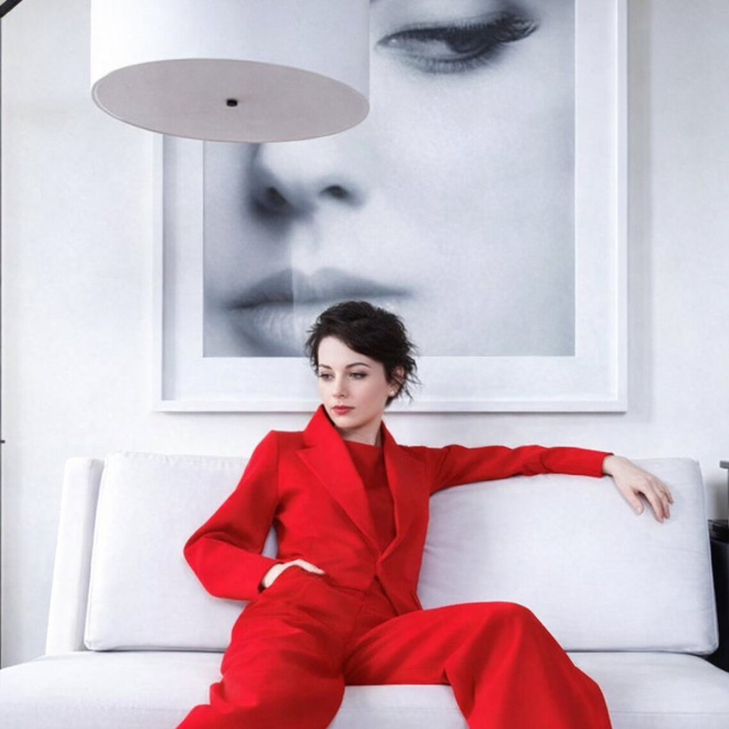 A woman with dark hair and fair skin sits confidently on a white sofa wearing a vivid red tailored suit. Behind her hangs a large black‑and‑white photograph of her face, creating a striking contrast between the monochrome background and her bold outfit. The minimalist interior features clean lines and a white pendant lamp overhead, emphasizing the modern, artistic atmosphere.