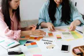 Two designers sit at a white table reviewing colour swatches, brand boards, and layout sketches, arranging palettes and notes as they plan a visual identity.