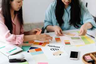 Two designers sit at a white table reviewing colour swatches, brand boards, and layout sketches, arranging palettes and notes as they plan a visual identity.