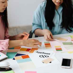 Two designers sit at a white table reviewing colour swatches, brand boards, and layout sketches, arranging palettes and notes as they plan a visual identity.