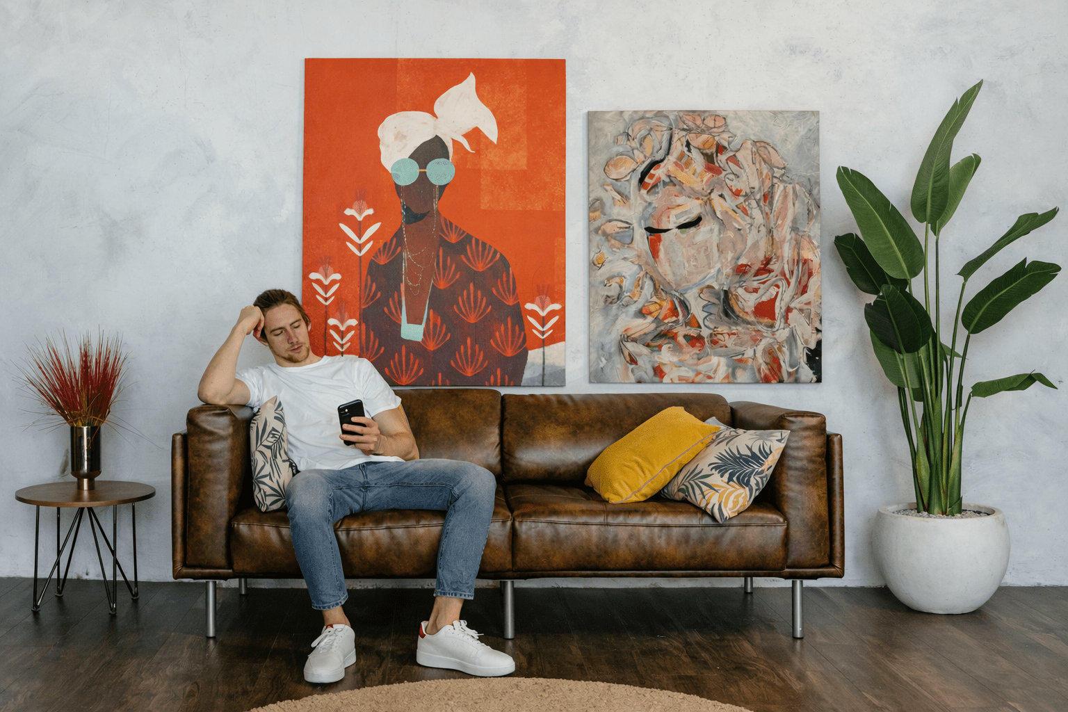 Young man sitting on a brown leather sofa in a modern living room with two expressive paintings on the wall — one bold orange figurative portrait and one abstract work titled Autumn Sylph, featuring gestural brushstrokes in white, pink, and ochre tones.