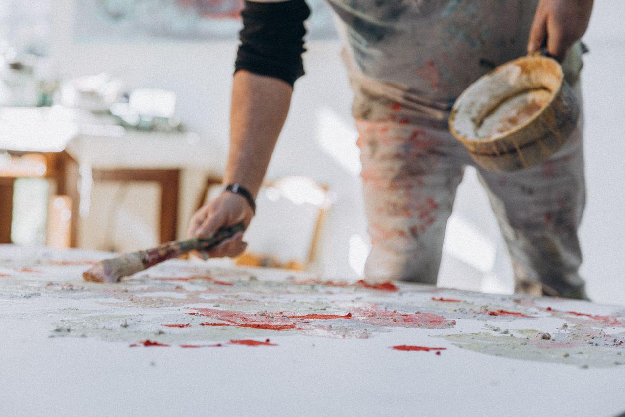 Artist applying thick paint layers to a large canvas in a bright studio, using a brush and bowl of pigment. The surface shows textured patches of red and pale tones, illustrating the tactile, process‑driven approach central to the Materiality and Surface Experimentation trend in contemporary Australian figurative painting.