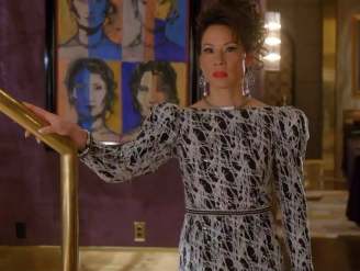 Lucy Liu as Simone Grove stands beside a gold staircase in Why Women Kill, wearing a black and white patterned gown. Behind her hangs a colourful pop‑art portrait painting by Spanish artist Manolo Valdés, featuring repeated faces in blue, orange and yellow tones against a deep purple wall.