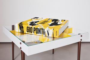 A large yellow art book titled Gio Ponti published by Taschen rests on a modern glass‑top coffee table. The book’s cover features bold black and white abstract brushstrokes, and its thick spine displays the title in black capital letters. The table has a white frame, brown legs, and geometric yellow panels beneath the glass, creating a clean, design‑focused composition that reflects Gio Ponti’s modernist aesthetic.