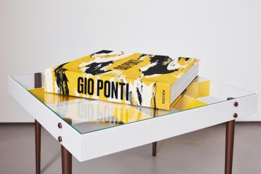 A large yellow art book titled Gio Ponti published by Taschen rests on a modern glass‑top coffee table. The book’s cover features bold black and white abstract brushstrokes, and its thick spine displays the title in black capital letters. The table has a white frame, brown legs, and geometric yellow panels beneath the glass, creating a clean, design‑focused composition that reflects Gio Ponti’s modernist aesthetic.