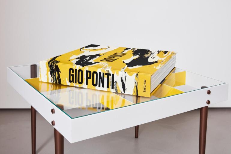 A large yellow art book titled Gio Ponti published by Taschen rests on a modern glass‑top coffee table. The book’s cover features bold black and white abstract brushstrokes, and its thick spine displays the title in black capital letters. The table has a white frame, brown legs, and geometric yellow panels beneath the glass, creating a clean, design‑focused composition that reflects Gio Ponti’s modernist aesthetic.