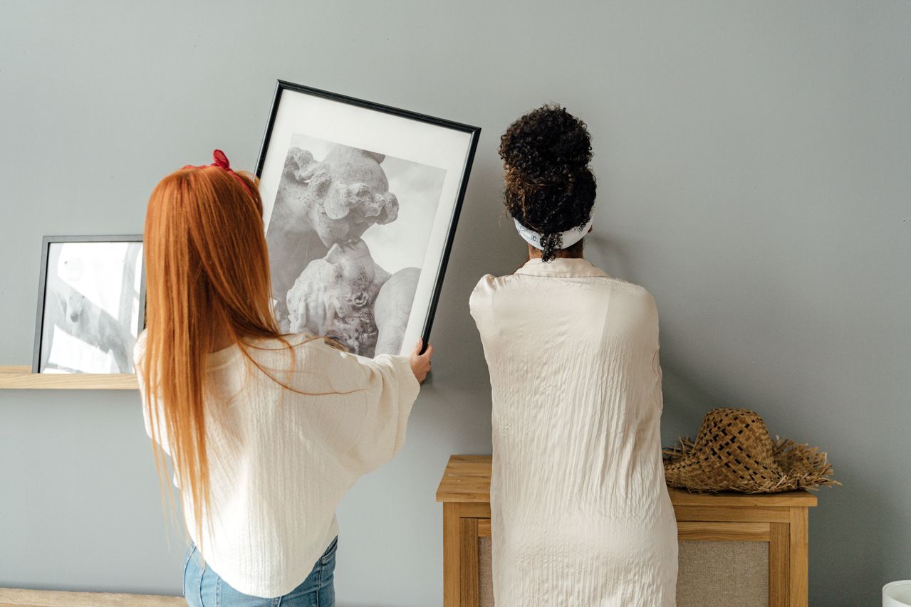 Two women seen from behind hanging framed monochrome artwork on a minimalist light grey wall. The woman on the left has long red hair and a cream knit sweater, carefully lifting a black-framed print depicting a classical marble figurative sculpture. The woman on the right has curly black hair tied in a high bun with a white headband, wearing a long beige textured linen shirt. A light wood storage cabinet sits against the wall beneath the artwork.