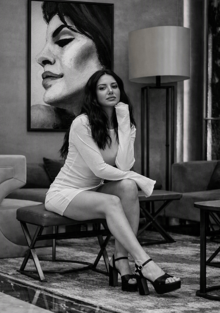 Black‑and‑white photograph of a woman seated on a cushioned bench in a modern lounge. She wears a long‑sleeved white mini dress and black platform heels, gazing directly at the camera with her chin resting on her hand. Behind her hangs a large framed ink drawing of a woman intertwined with leaves, rendered in expressive monochrome lines. The composition balances elegance and contrast, blending contemporary interior design with fine‑art atmosphere.