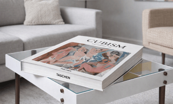 Cubism art book by Anne Ganteführer‑Trier displayed on a modern glass‑topped coffee table in a minimalist living room. The book’s cover shows Picasso’s Les Demoiselles d’Avignon with angular pink and blue figures. A light grey sofa, neutral rug, and soft lighting create a refined, contemporary setting.