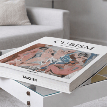 Cubism art book by Anne Ganteführer‑Trier displayed on a modern glass‑topped coffee table in a minimalist living room. The book’s cover shows Picasso’s Les Demoiselles d’Avignon with angular pink and blue figures. A light grey sofa, neutral rug, and soft lighting create a refined, contemporary setting.