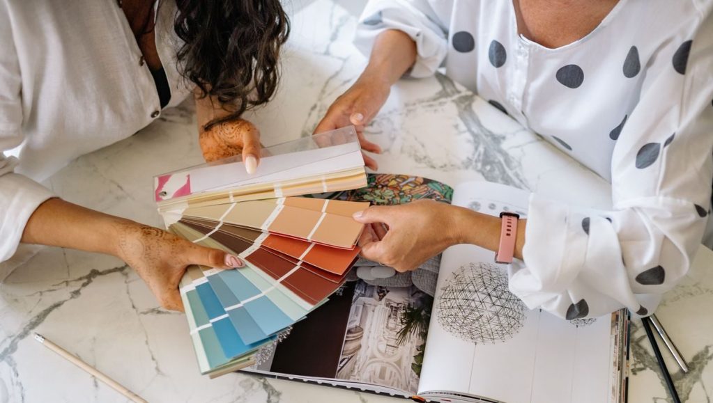 Two people seated at a marble table reviewing a fan deck of colour swatches ranging from blues to warm neutrals, alongside an open interior design book showing room photography and lighting sketches — a collaborative moment of design planning and palette selection.