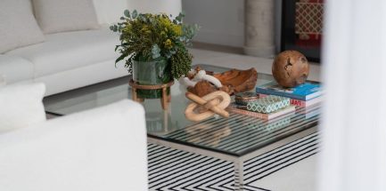 Contemporary living room with a glass coffee table styled with natural wood decor, art books, and a lush green floral arrangement in a glass vase; surrounded by white sofas and a geometric black‑and‑white rug for a refined, organic aesthetic.