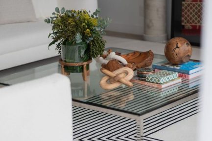 Contemporary living room with a glass coffee table styled with natural wood decor, art books, and a lush green floral arrangement in a glass vase; surrounded by white sofas and a geometric black‑and‑white rug for a refined, organic aesthetic.