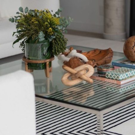 Contemporary living room with a glass coffee table styled with natural wood decor, art books, and a lush green floral arrangement in a glass vase; surrounded by white sofas and a geometric black‑and‑white rug for a refined, organic aesthetic.