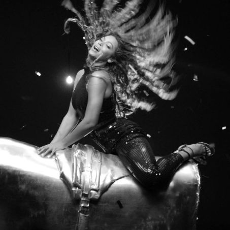 Black‑and‑white photograph of Beyoncé in motion, captured mid‑performance on a large metallic cylinder. Her sequined outfit catches the light as her hair fans outward dramatically, creating a sense of rhythm and power. The industrial backdrop and reflective surfaces evoke the *Suga Mama* video’s monochrome aesthetic — a fusion of sensual movement, texture, and strength that defines the Sonic Noir style.